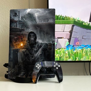 May include: A black PlayStation 5 console with a custom skin featuring a man in a gas mask sitting on a chair in front of a building with the word "Припять" written on it. The console is on a white surface with a black PlayStation 5 controller next to it.
