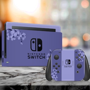 Pode incluir: Um conjunto de consola e comando Nintendo Switch roxo. A consola e o comando são decorados com um design floral. O logótipo Nintendo Switch é visível tanto na consola como no comando.