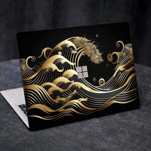 May include: Laptop with a black cover featuring a gold wave design. The wave pattern is stylized with swirling lines and intricate details. The laptop is open, revealing the keyboard and screen.