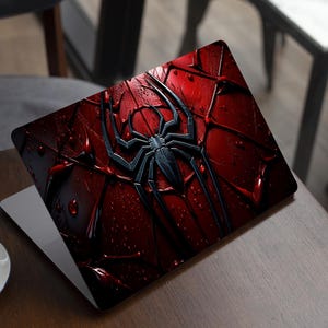 May include: Laptop skin with a black spider emblem on a textured, deep red background. The design has a 3D effect with water droplet details. The laptop is open on a wooden table, with a coffee cup in the foreground.