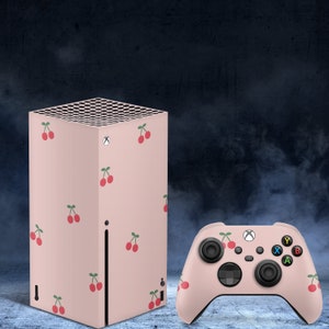 May include: A pink Xbox Series X console with a cherry pattern and a matching pink Xbox controller. The console has a black front panel with a white Xbox logo.