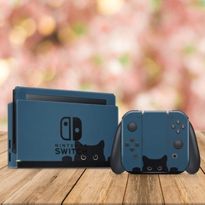 Peut inclure: Une console Nintendo Switch bleue et des manettes Joy-Con assorties avec un motif de chat noir. La console affiche le logo Nintendo Switch et le texte "NINTENDO SWITCH". Les manettes Joy-Con ont un chat noir qui se cache derrière les boutons.