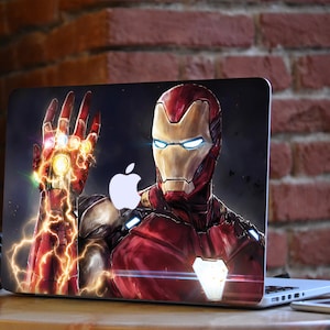 May include: A laptop computer with a custom design featuring Iron Man from the Marvel Cinematic Universe. The design shows Iron Man with his gauntlet raised, displaying the Infinity Stones. The laptop has a silver Apple logo on the lid.