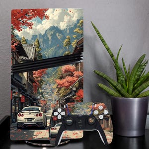 May include: A games console and controller with a Japanese street scene design, featuring red autumn leaves and a white sports car. The controller is black and white with matching artwork.