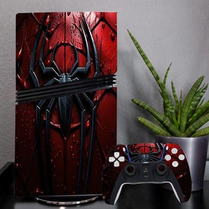 May include: A red and black gaming console with a matching controller. The console features a large spider graphic. The controller has a black base with red accents and white buttons. A potted plant sits to the right.
