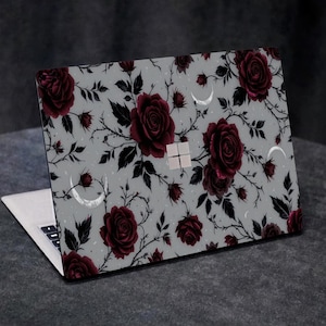 Peut inclure: Ordinateur portable avec un motif floral de roses rouge foncé, de feuilles noires et de croissants de lune sur un fond gris clair. L'ordinateur portable est ouvert, révélant le clavier et l'écran. Le motif recouvre le dessus.