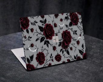 May include: Laptop with a floral design featuring dark red roses, black leaves, and crescent moons on a light gray background. The laptop is open, revealing the keyboard and screen. The design covers the top of the laptop.