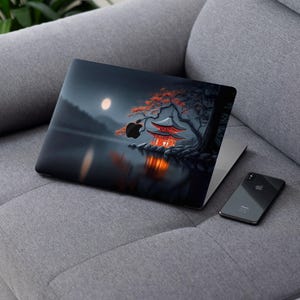 May include: Laptop computer with a scenic Japanese-inspired design featuring a pagoda, a full moon, and a tree with red leaves. A black smartphone rests beside the laptop on a gray sofa.