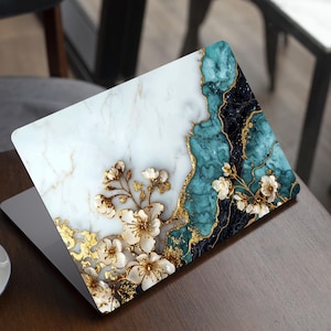 May include: Laptop with a decorative cover. The cover has a marble design with teal, black, and gold accents. White floral embellishments and gold leaf details adorn the cover, creating an elegant aesthetic. The laptop sits on a wooden table.