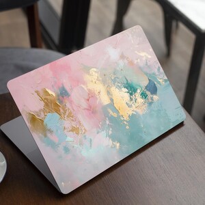 May include: Laptop with an abstract art design. The cover displays a mix of pink, gold, teal, and blue brushstrokes, giving a textured, artistic appearance. The laptop is open on a wooden table, with a coffee cup in the foreground.
