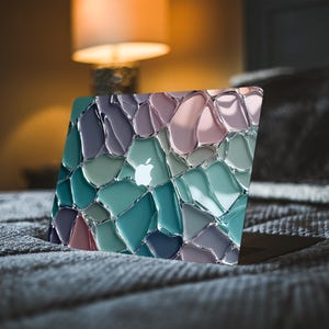 May include: A laptop with a colorful, mosaic-style design on the lid. The design features irregular shapes in shades of blue, green, purple, and pink, with a silver Apple logo. The laptop is resting on a gray, textured surface.
