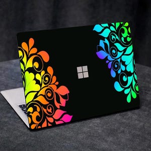 Può includere: Laptop nero con un colorato motivo floreale sul coperchio. Il design presenta una sfumatura di colori, tra cui arancione, giallo, verde, blu e viola. Il laptop ha una tastiera argentata e un logo Microsoft.
