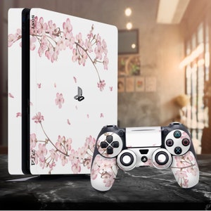 Peut inclure: Une console PlayStation 4 blanche avec un motif de cerisier rose et une manette assortie. La console porte l'inscription "SONY" et "PS4".