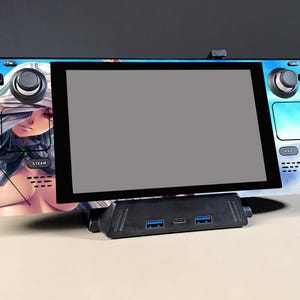 May include: A Steam Deck handheld gaming console with a custom skin featuring an anime-style illustration. The console is docked on a black hub with USB ports. The screen is gray.