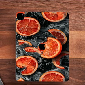 May include: A tablet skin featuring a vibrant pattern of sliced blood oranges set against a dark, swirling liquid background. The design incorporates a black Apple logo cutout positioned centrally, surrounded by bright orange citrus segments.