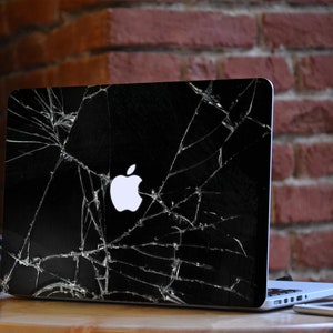May include: A black laptop with a cracked screen and a white Apple logo. The laptop is sitting on a wooden table with a pair of headphones and a smartphone next to it.