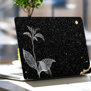 May include: A black laptop with a white floral design and gold glitter accents on the lid. The laptop is closed and sitting on a white table.