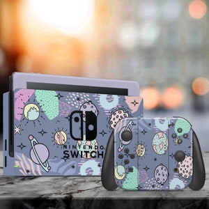 Può includere: Una skin per console e controller Nintendo Switch di colore viola e grigio con un design a tema spaziale che presenta pianeti, stelle e forme geometriche. Il testo "NINTENDO SWITCH" è stampato sulla skin della console.