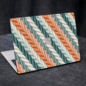 Può includere: Laptop con un design a treccia colorato. Il motivo presenta strisce diagonali arancioni, bianche e verde acqua. Il laptop è aperto, mostrando la tastiera e un logo argentato.