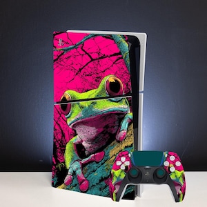 Op de afbeelding: Een spelcomputer en bijpassende draadloze controller met een levendige grafische skin. Het ontwerp toont een felgroene kikker met grote rode ogen op een getextureerde tak tegen een felroze achtergrond met zwarte silhouetten van bomen.