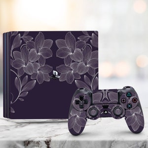 Könnte beinhalten: Eine schwarze PlayStation 4 Konsole und ein Controller mit einem weißen Blumenmuster. Die Konsole hat ein elegantes, modernes Design und der Controller liegt angenehm in der Hand.