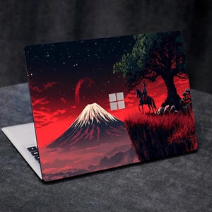 Op de afbeelding: Laptop met een aangepaste skin met een dramatisch landschap. Het ontwerp omvat een besneeuwde berg, een ruiter te paard en een grote boom tegen een rode hemel met sterren. Het Windows-logo is zichtbaar.