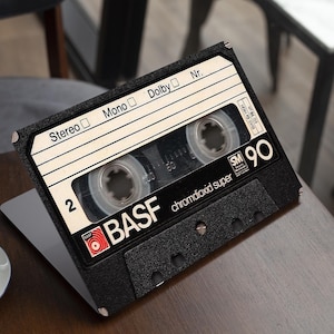 Peut inclure: Une cassette noire avec des détails blancs et rouges, marquée "BASF chromdioxid super SM 90". La cassette est posée sur un ordinateur portable sur une table en bois, avec une tasse de café au premier plan. La cassette porte les mots "Stereo", "Mono" et "Dolby".