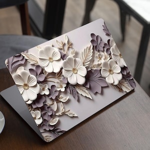 Può includere: Laptop con un design floreale. La cover del laptop presenta un rilievo 3D di fiori bianchi e viola con accenti dorati e foglie. Il laptop è aperto su un tavolo di legno, con una tazza di caffè in primo piano.