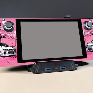 Puede incluir: Consola de juegos portátil rosa con pantalla negra, con un diseño de coche y flores de cerezo. La consola descansa sobre una estación de acoplamiento negra. El diseño incluye una cruceta, joysticks y botones.