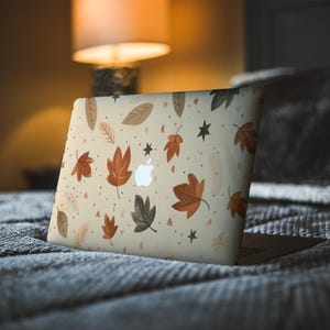 May include: A laptop with a beige cover featuring a fall-themed design. The cover is adorned with illustrations of autumn leaves in shades of brown, orange, and green, along with small black stars. The Apple logo is visible.