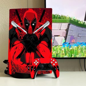 Könnte beinhalten: Eine rote und schwarze PlayStation 5 Konsole und ein Controller mit Deadpool-Design. Die Konsole hat einen roten und schwarzen Hintergrund mit einem Deadpool-Charakter in der Mitte. Der Controller hat ein rotes und schwarzes Design mit Deadpools Augen auf der Vorderseite.