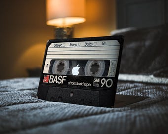 May include: A black BASF chromdioxid super 90 audio cassette tape with a white Apple logo. The cassette is on a grey textured surface. The tape has white and black labels with text and a number 2.