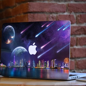 May include: A laptop with a colorful galaxy and cityscape design. The design features a purple sky with shooting stars, a cityscape with skyscrapers, and a planet in the background. The laptop has a silver Apple logo on the lid.