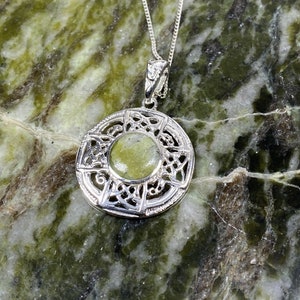 Pode incluir: Um colar de prata com um pingente de nó celta com uma pedra verde no centro. O pingente é redondo e tem um design detalhado. O colar está sobre um fundo de mármore verde e branco.