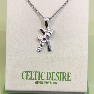 Può includere: Collana con pendente in argento a forma di lettera "K" con una piccola gemma rossa. Il pendente è appeso a una delicata catena ed è esposto su una carta bianca con il testo "Celtic Desire Silver Jewellery".