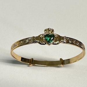 May include: A gold-toned bangle bracelet featuring a Claddagh design at the center. The centerpiece includes two hands holding a heart-shaped green gemstone topped with a crown. The band is engraved with intricate scrollwork patterns.