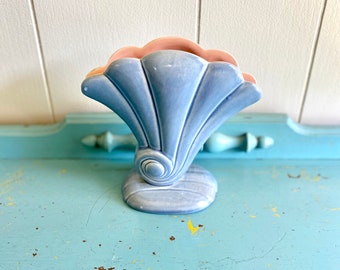 May include: A light blue ceramic vase with a pink interior and a shell-like design. The vase is sitting on a blue and yellow painted surface.