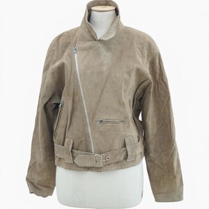 May include: A tan suede jacket with an asymmetrical zipper closure and a notched collar. The jacket features a small zippered pocket on the chest and a belt detail at the waist. The jacket is displayed on a mannequin.