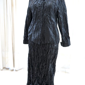 May include: A black, long-sleeved shirt and skirt set made of a crinkled fabric. The shirt has a button-up front and a collar. The skirt is long and flowing.