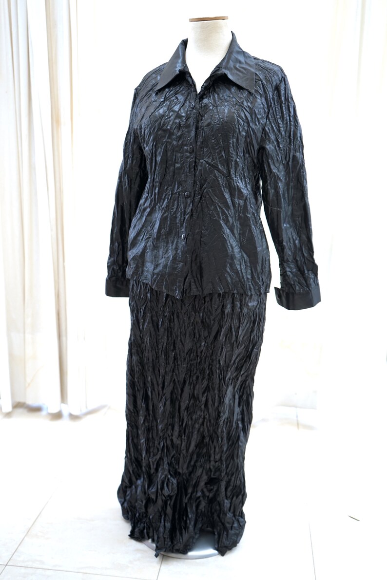 May include: A black, long-sleeved shirt and skirt set made of a crinkled fabric. The shirt has a button-up front and a collar. The skirt is long and flowing.