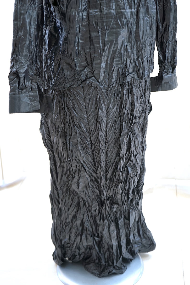 May include: A black long-sleeved shirt with a crinkled texture. The shirt has a collar and long sleeves with button cuffs. The shirt is made of a soft, flowing fabric.