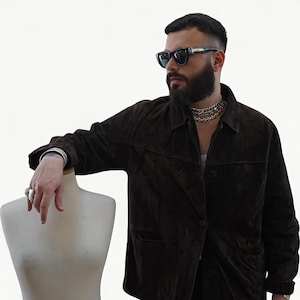 May include: A dark brown suede jacket with a button-up front and two front pockets. The jacket is worn by a person wearing sunglasses and a silver chain necklace. A silver ring and bracelet are also visible.