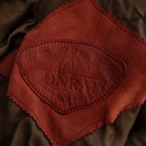 Puede incluir: Primer plano de un parche de cuero marr&oacute;n con un borde cosido. El parche presenta un logotipo en relieve, estampado, que dice "ONRAI" con un dise&ntilde;o estilizado.