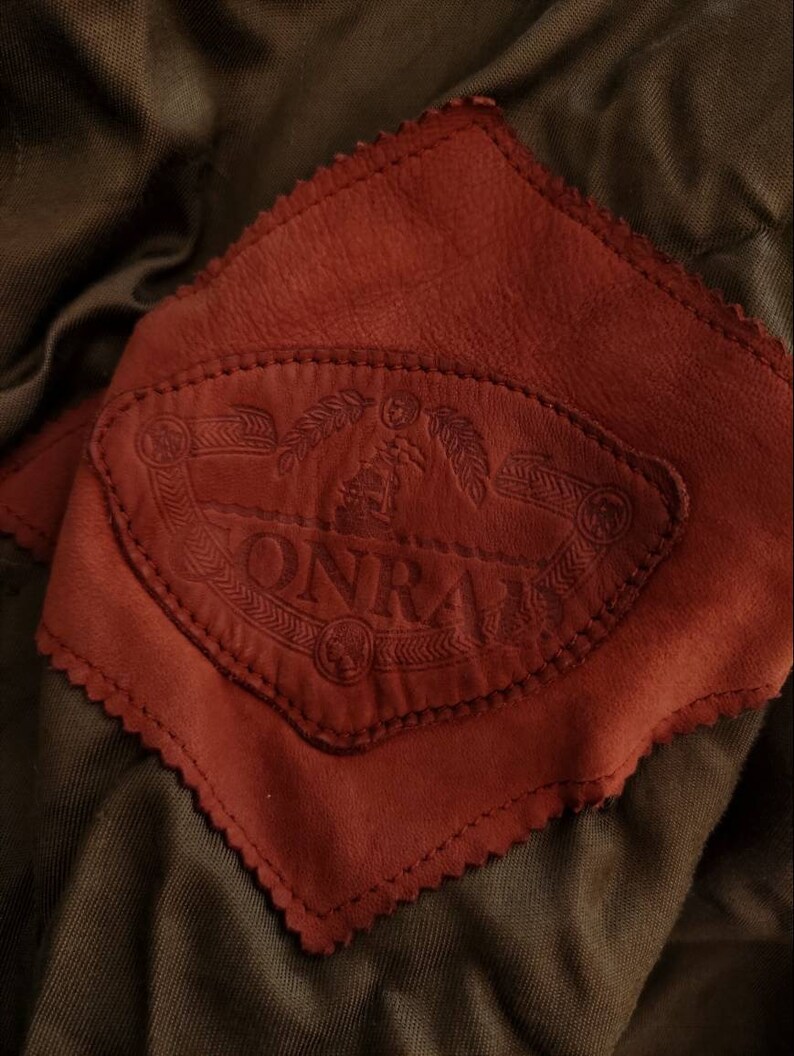 Puede incluir: Primer plano de un parche de cuero marr&oacute;n con un borde cosido. El parche presenta un logotipo en relieve, estampado, que dice "ONRAI" con un dise&ntilde;o estilizado.