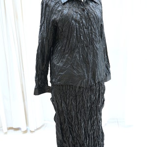 May include: A black, long-sleeved shirt and skirt set made of crinkled fabric. The shirt has a collar and button closure. The skirt is long and flowing.