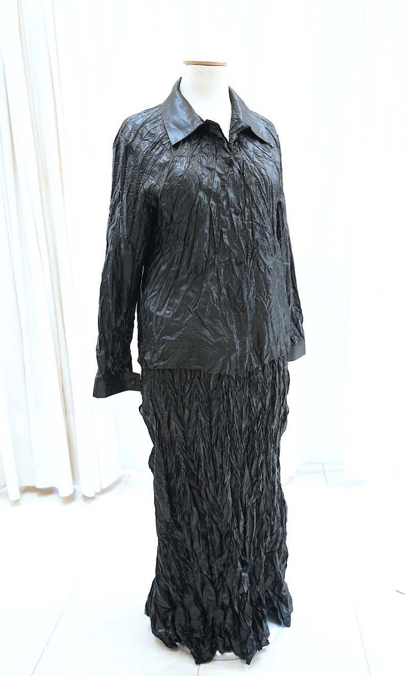 May include: A black, long-sleeved shirt and skirt set made of crinkled fabric. The shirt has a collar and button closure. The skirt is long and flowing.