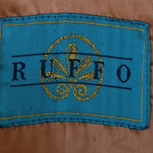 K&ouml;nnte beinhalten: Ein t&uuml;rkisfarbenes Stofflabel mit einem gelben, gesteppten Rand. Das Label tr&auml;gt den Schriftzug "RUFFO" in schwarzer Schrift und ein goldfarbenes, gesticktes Blumenmuster.