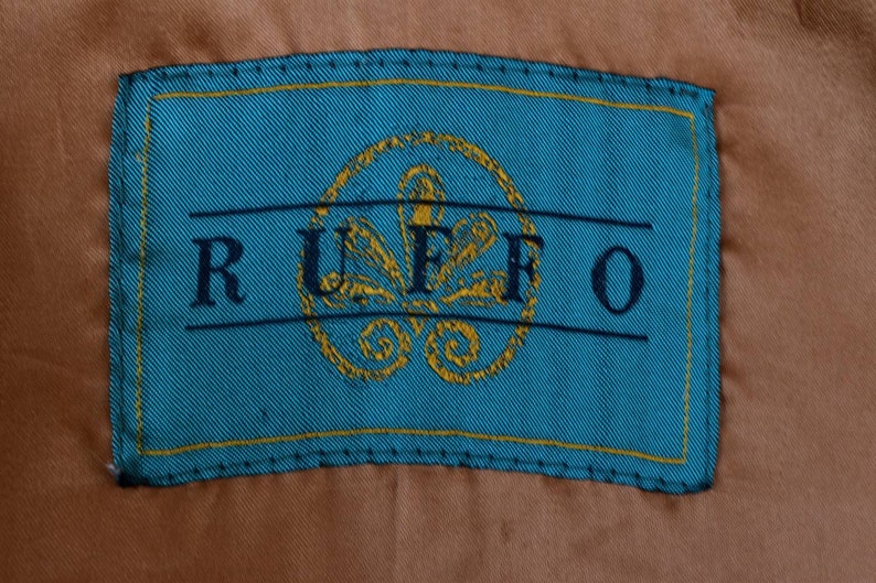 K&ouml;nnte beinhalten: Ein t&uuml;rkisfarbenes Stofflabel mit einem gelben, gesteppten Rand. Das Label tr&auml;gt den Schriftzug "RUFFO" in schwarzer Schrift und ein goldfarbenes, gesticktes Blumenmuster.