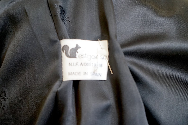 K&ouml;nnte beinhalten: Nahaufnahme eines schwarzen Stoffes mit einem Etikett. Das Etikett zeigt ein Eichh&ouml;rnchen-Logo und den Text "estgar sa", sowie "N.I.F. A08512118" und "MADE IN SPAIN".
