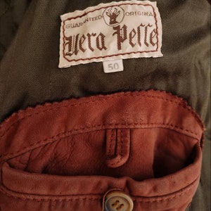 Puede incluir: Un bolsillo de parche de cuero marr&oacute;n con un bot&oacute;n en una chaqueta verde. La chaqueta tiene una etiqueta que dice "Guaranteed Original Vera Pelle" y "50".
